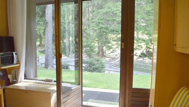 A room with a large sliding glass door leading to a forested area, a small wooden table with a microwave, and a striped chair.