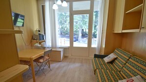A room with a bed, a desk, and a chair. There is a window with a view of trees and a car outside.