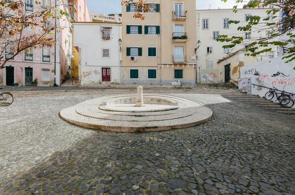 A cobblestone courtyard with a central fountain, surrounded by multi-story buildings with balconies and windows.