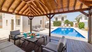 An outdoor pool area with a covered patio, lounge chairs, and a dining table set for two.