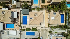 An aerial view of a resort with multiple swimming pools, some with sun loungers, surrounded by palm trees and a tiled roof structure.