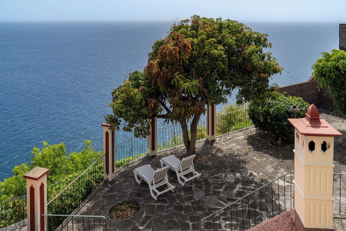 A terrace with stone flooring, two white lounge chairs, a tree, and a view of the ocean.