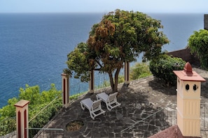 Una terraza con suelo de piedra, dos sillones blancos, un árbol y vista al océano.