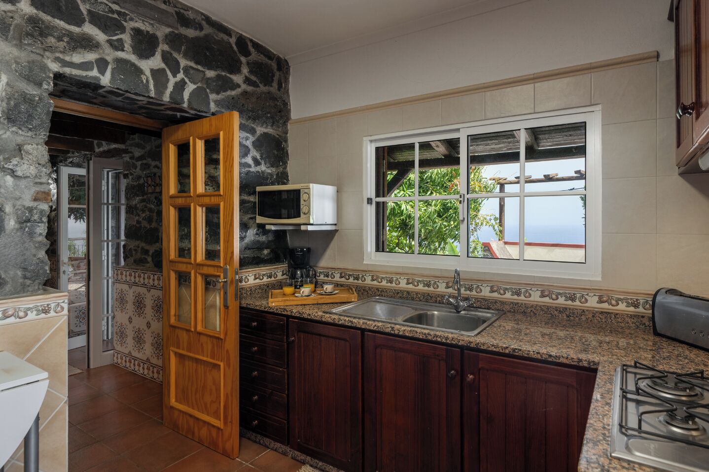 A kitchen with a stone wall, wooden cabinets, a microwave, a sink, and a window with a view.