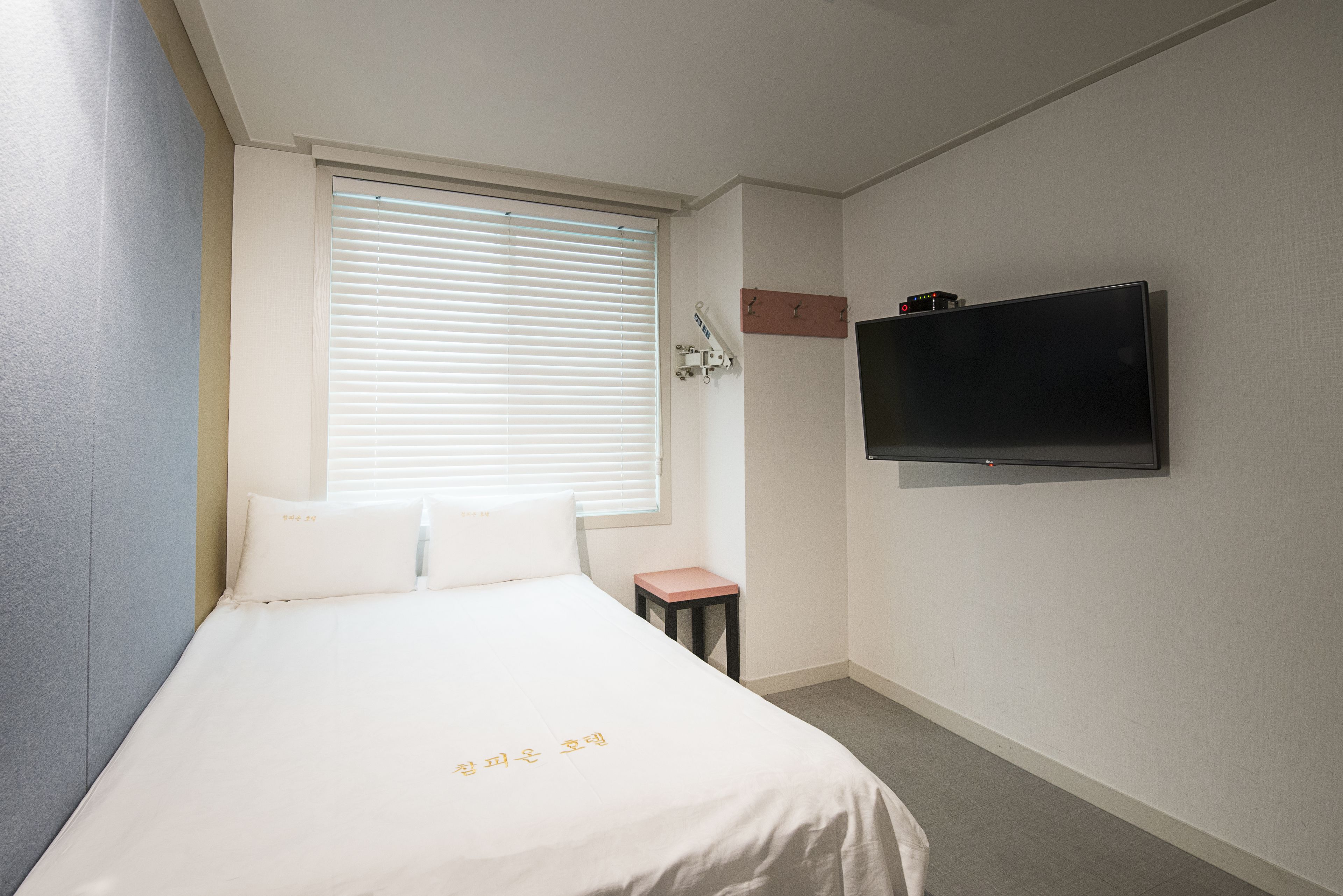 A hotel room with a bed, a small table, a TV mounted on the wall, and a window with blinds.