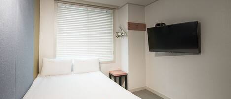 A hotel room with a bed, a small table, a TV mounted on the wall, and a window with blinds.