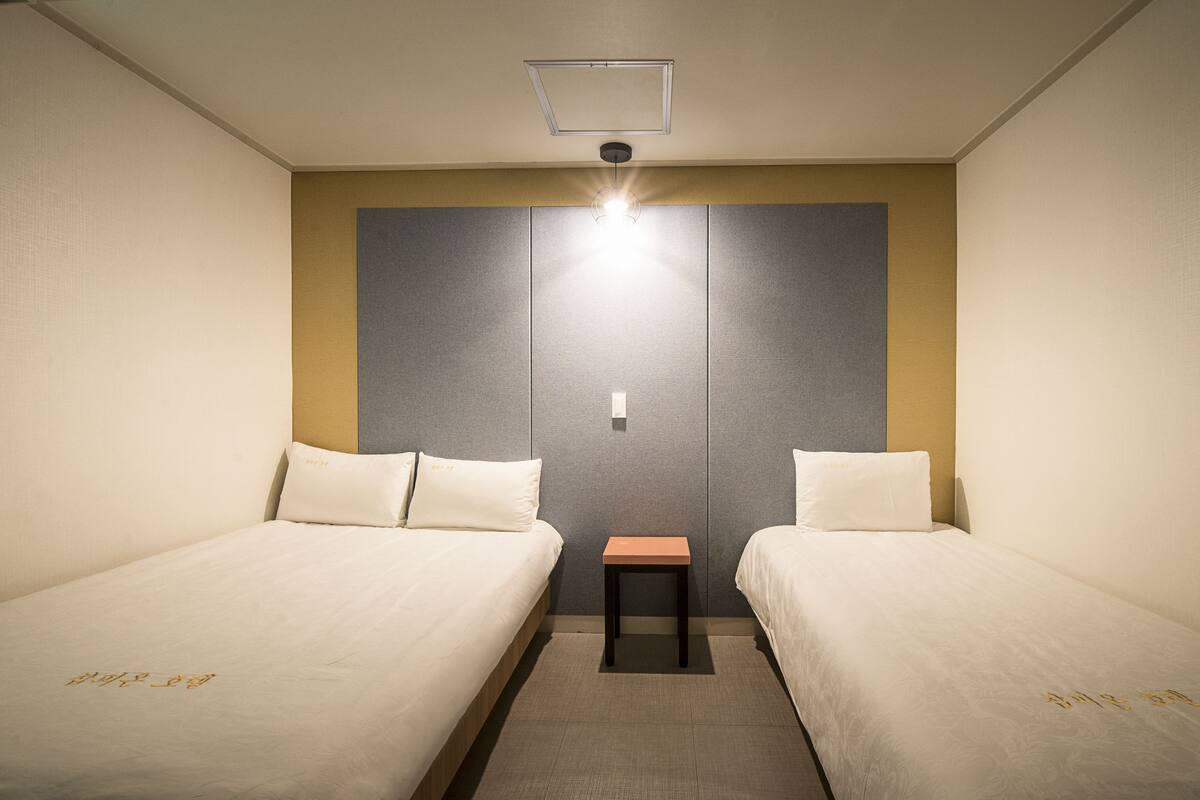 A hotel room with two single beds, a small table, and a light fixture above the bed.