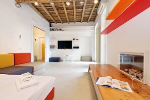 A modern hotel room with a wooden ceiling, a flat-screen TV, a sofa, a dining table, and a fireplace.