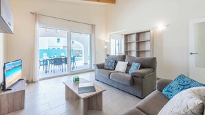 En moderne stue med sofa, sofabord og fjernsyn. Der er en skydedør til et udendørs område med pool og stole.