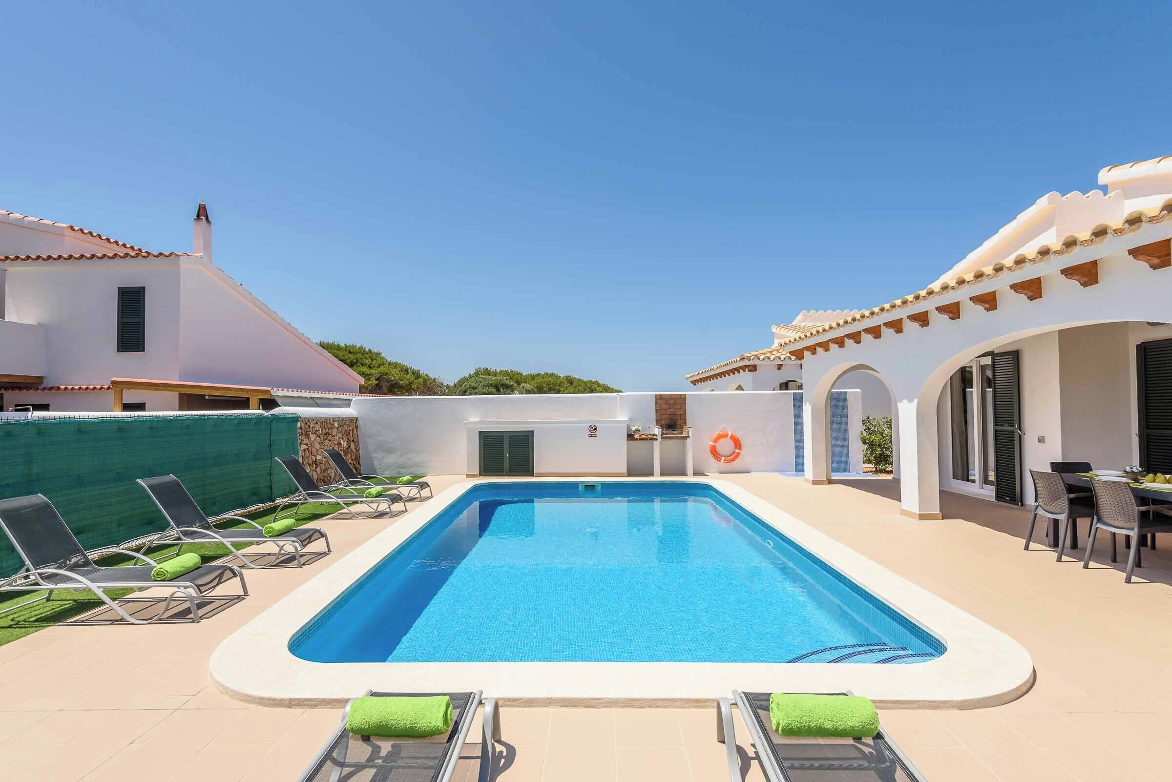 Una piscina rectangular con un solárium empedrado, rodeada de sillones de descanso y un área de patio cubierta.