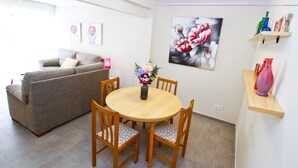 A living room with a grey sofa, a wooden dining table with four chairs, and a vase of flowers. There are two framed floral paintings on the wall, a shelf with bottles, and a pink vase on a wooden shelf.