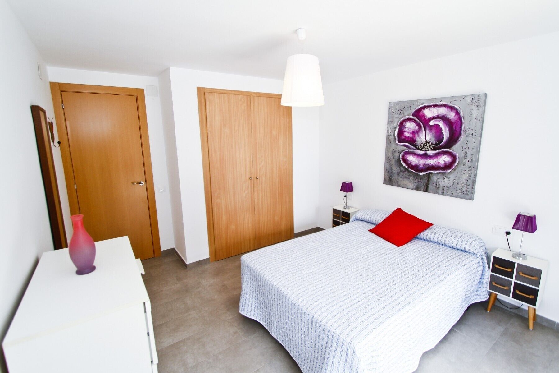 A bedroom with a bed, a bedside table, a lamp, a painting, and a wardrobe.