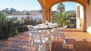 A terrace with a table and chairs, a bottle of wine, and a view of a white building and palm trees.