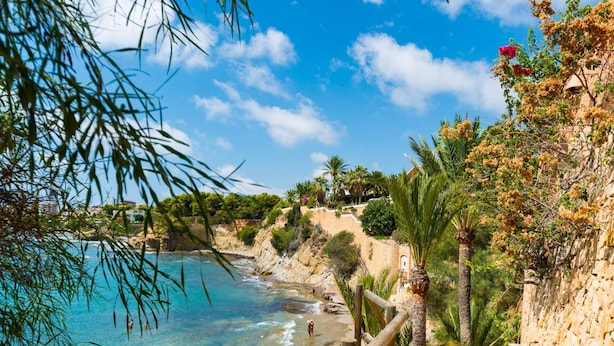 A coastal view with clear turquoise waters, a stone staircase leading down to the beach, and lush greenery.