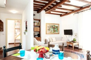 A living room with a wooden ceiling, a sofa, a TV, a bookshelf, and a dining area with a table set for two.