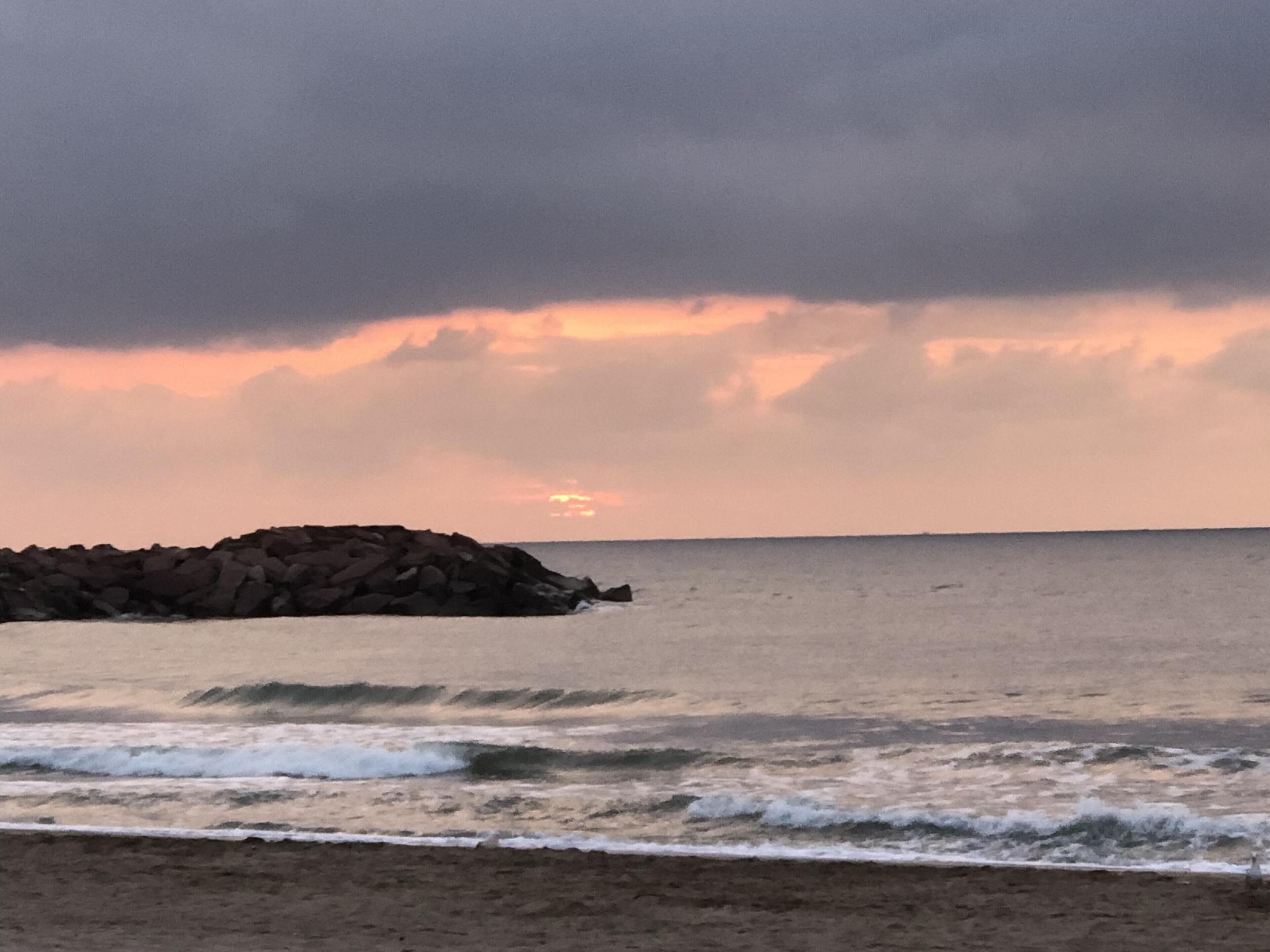 A beach with waves, a rocky outcrop, and a sunset.