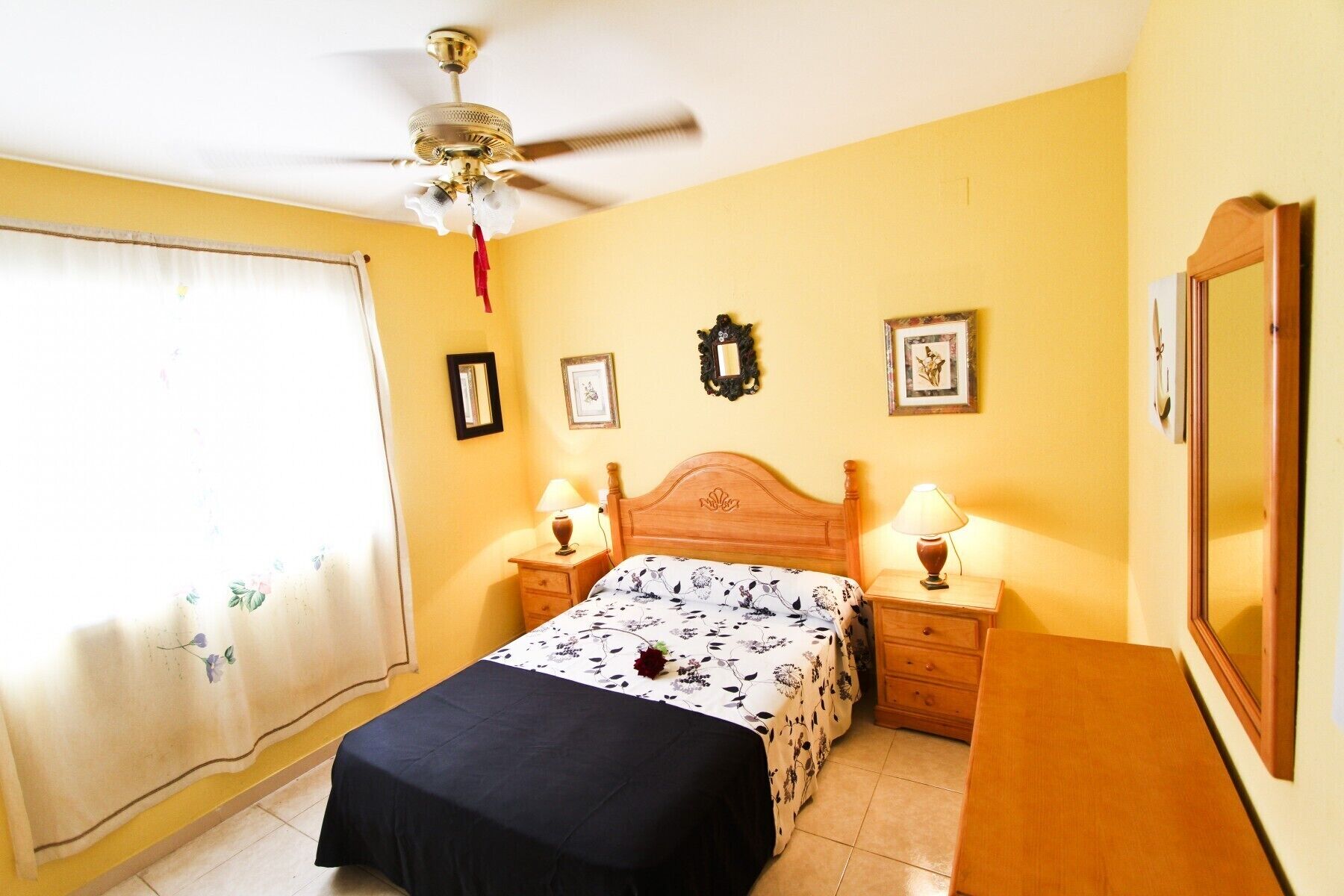 A bedroom with a bed, two bedside tables with lamps, a ceiling fan, and wall-mounted pictures.