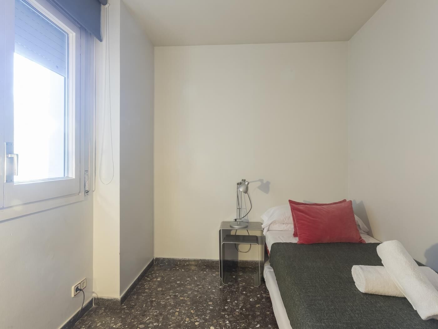 A small, clean room with a bed, a nightstand, a lamp, and a window.