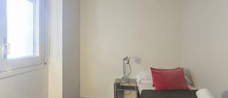 Ein kleiner, sauberer Raum mit einem Bett, einem Nachttisch, einer Lampe und einem Fenster.