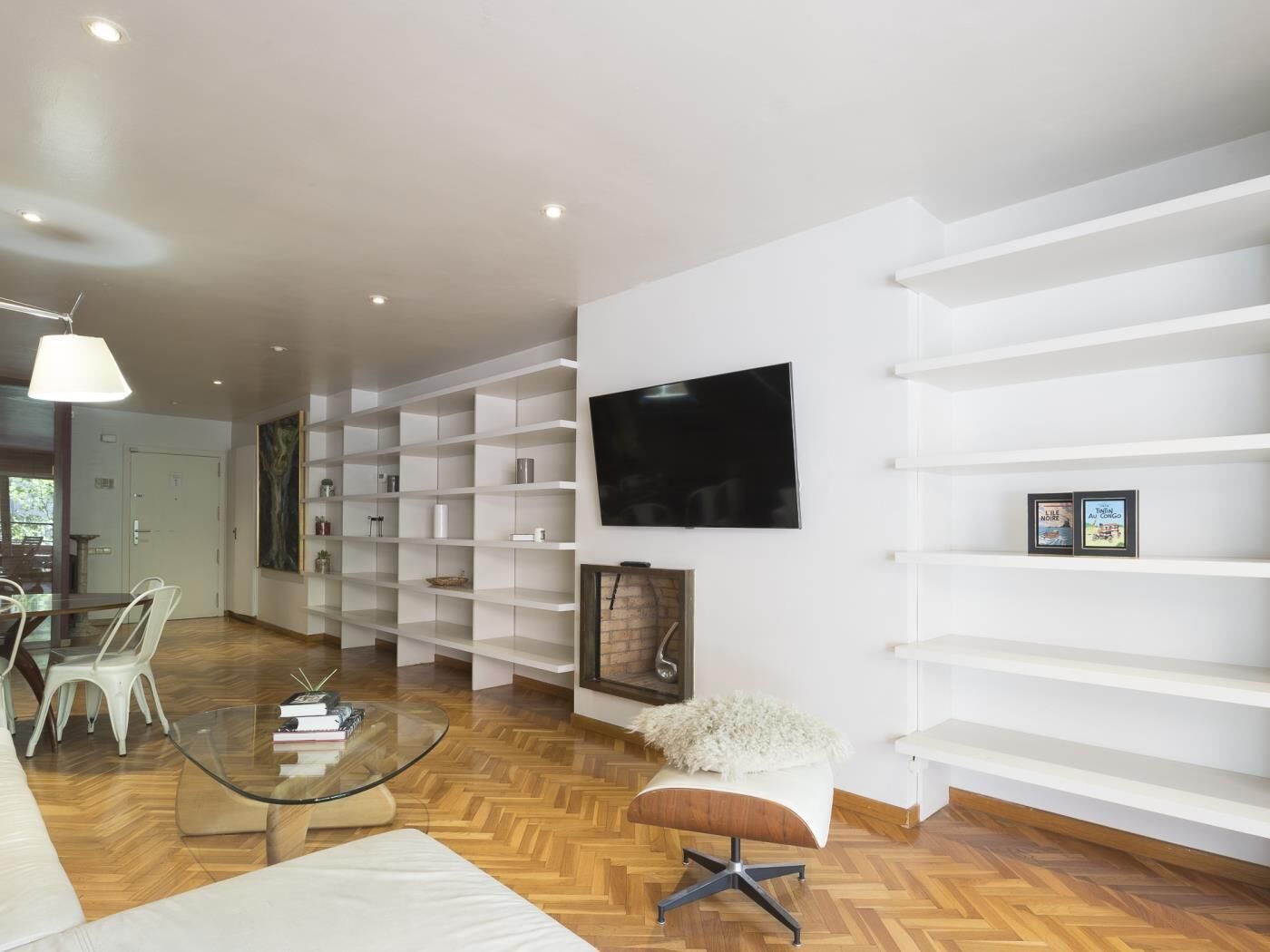 Een moderne woonkamer met een glazen salontafel, een witte bank en een witte poef. Er staan witte planken langs de wanden en een flatscreen televisie is daarboven bevestigd.