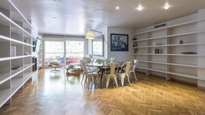A spacious room with a large dining table, white chairs, and a herringbone wood floor. There are white shelving units on both sides of the room and a large window providing natural light. - My Space Barcelona apartment very close to Teknon Medical Center (Barcelona)