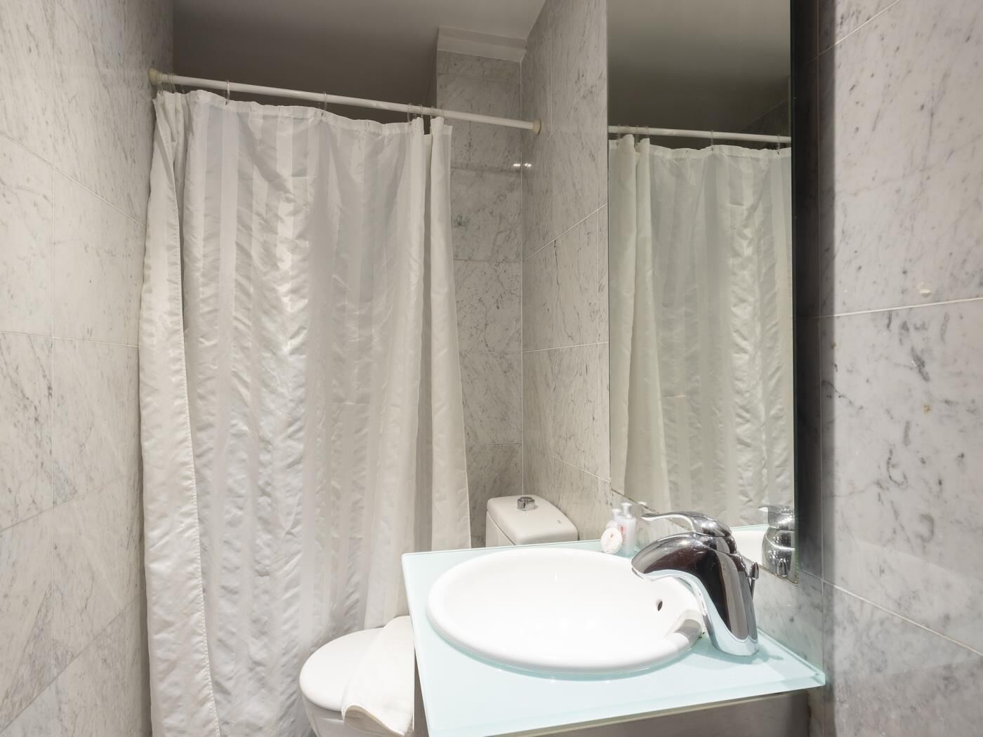 Un baño con inodoro blanco, lavamanos de vidrio y cortina de ducha.