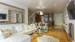 A modern living room with a white sofa, a glass coffee table, and a large mirror. There are shelves with decorative items and a television mounted on the wall. - My Space Barcelona apartment very close to Teknon Medical Center (Barcelona)