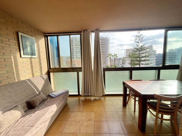 A living room with a sofa, a dining table with chairs, a large window with curtains, and a view of buildings and palm trees. - ALBAIDA PARK BLUELINE R014 (Benidorm)