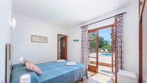 Quarto com cama, mesa de cabeceira com luminária, outra mesa de cabeceira menor com luminária, janela com cortinas e vista para uma piscina.