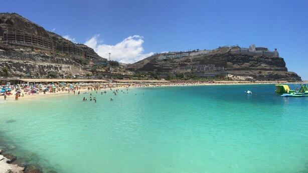 Une plage à l’eau turquoise et limpide, un rivage sablonneux et, au loin, sur une colline, un grand bâtiment.