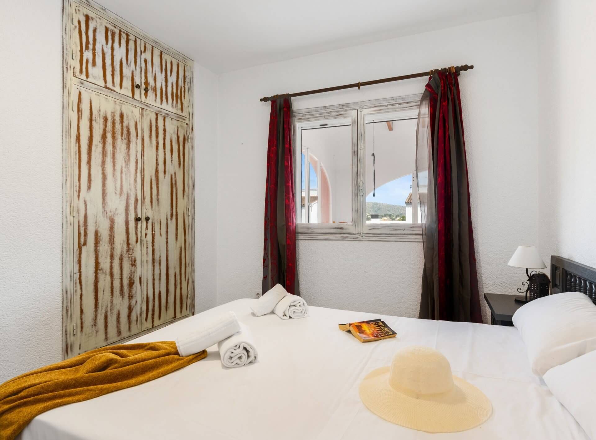 A hotel room with a bed, a window with red curtains, a bedside table with a lamp, and a view of the outside.
