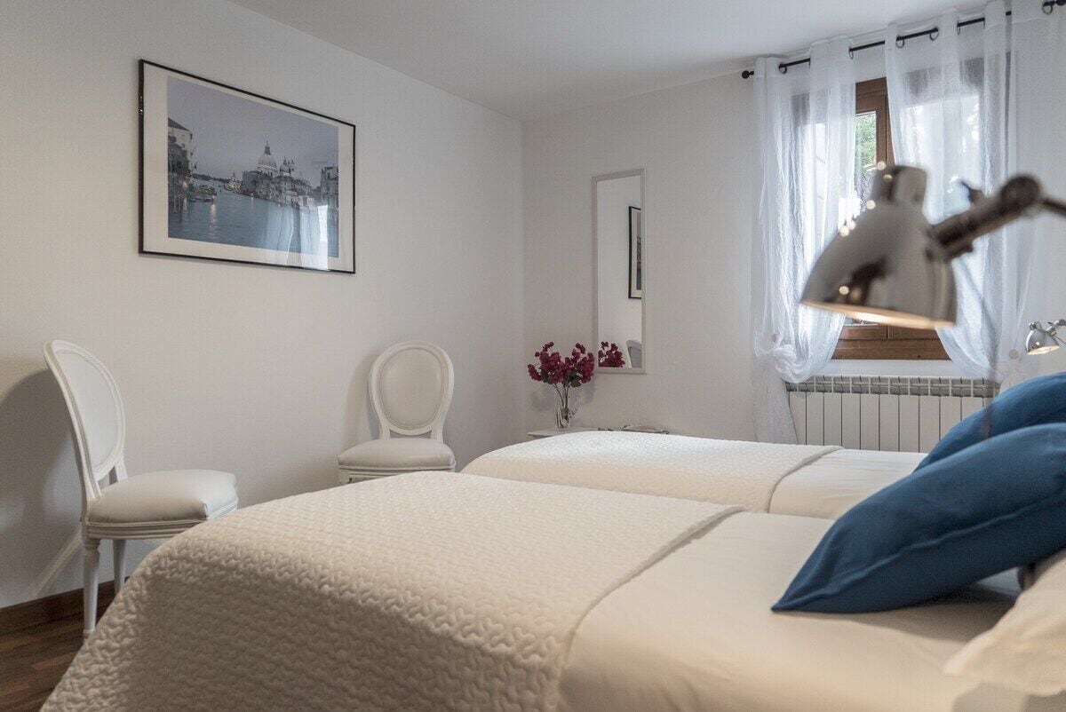 Habitación de hotel con dos camas, una silla, una mesa, un cuadro en la pared y una ventana con cortinas.