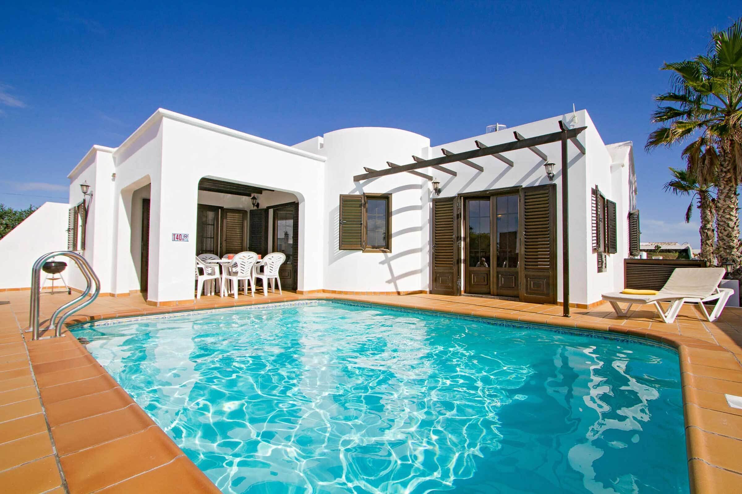 Ein Poolbereich mit klarem, blauem Pool, einem weißen Gebäude mit einer Pergola und einer Palme.