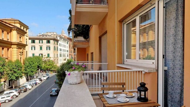 A balcony with a table, chairs, a basket of flowers, and a view of a street with parked cars and buildings.