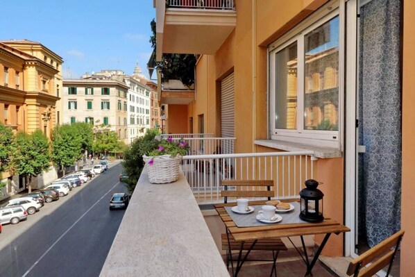 A balcony with a table, chairs, a basket of flowers, and a view of a street with parked cars and buildings.