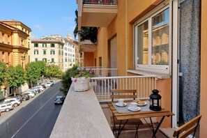Un balcon avec une table, des chaises, un panier de fleurs et une vue sur une rue bordée de voitures garées et d’immeubles.