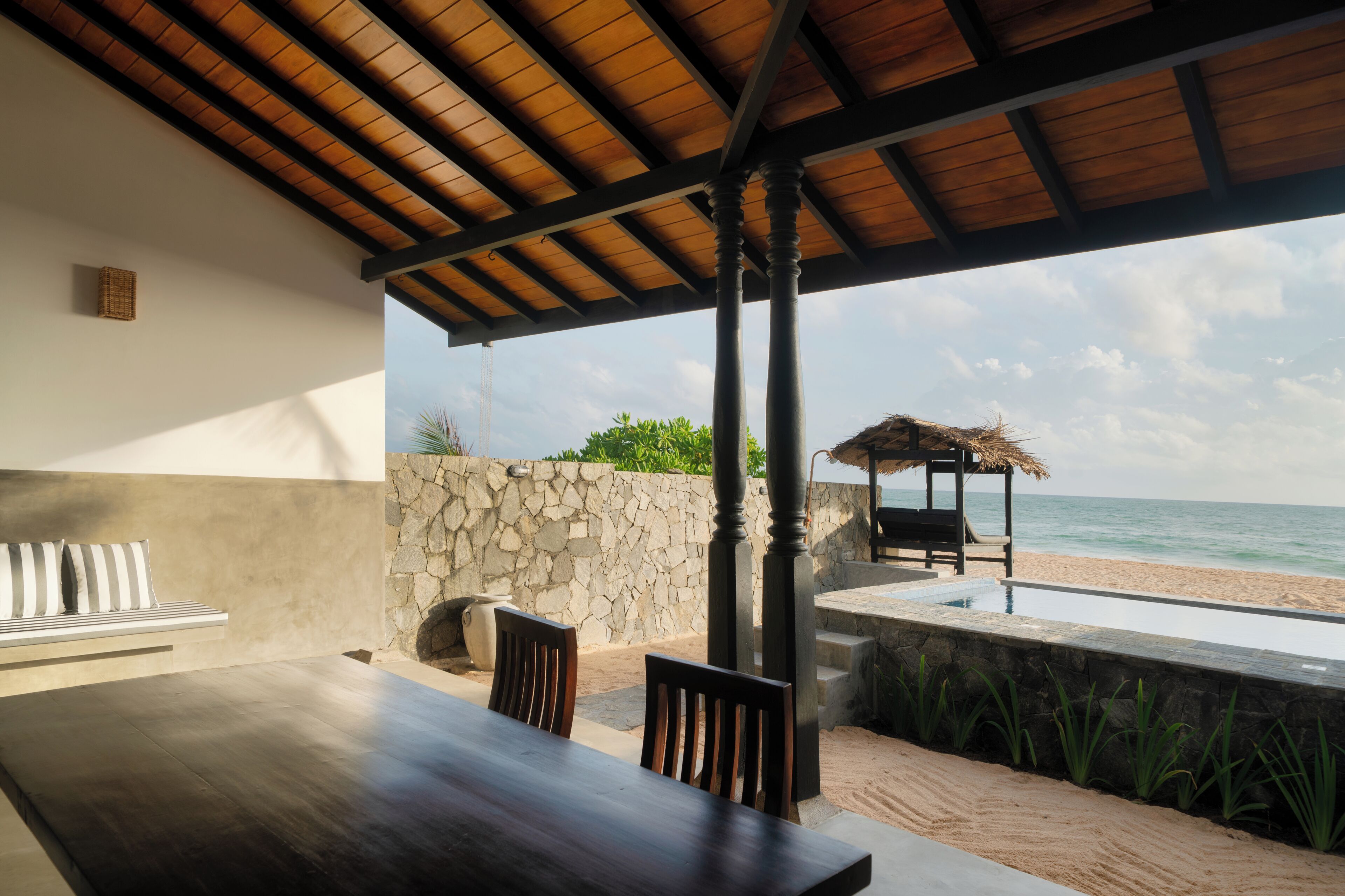 An outdoor seating area with a table and chairs, a stone wall, and a view of the beach and ocean.