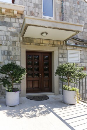 A stone building with a brown door, two potted plants, and a small awning above the door.