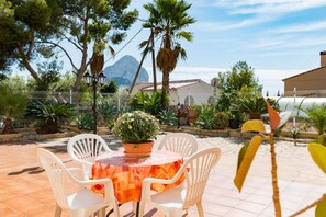 Une terrasse extérieure avec une table ronde et des chaises blanches, entourée de plantes en pot et offrant une vue sur une montagne.