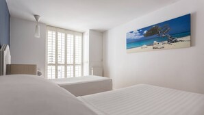 A hotel room with two beds, a window with blinds, a painting of a beach scene, and a ceiling light.