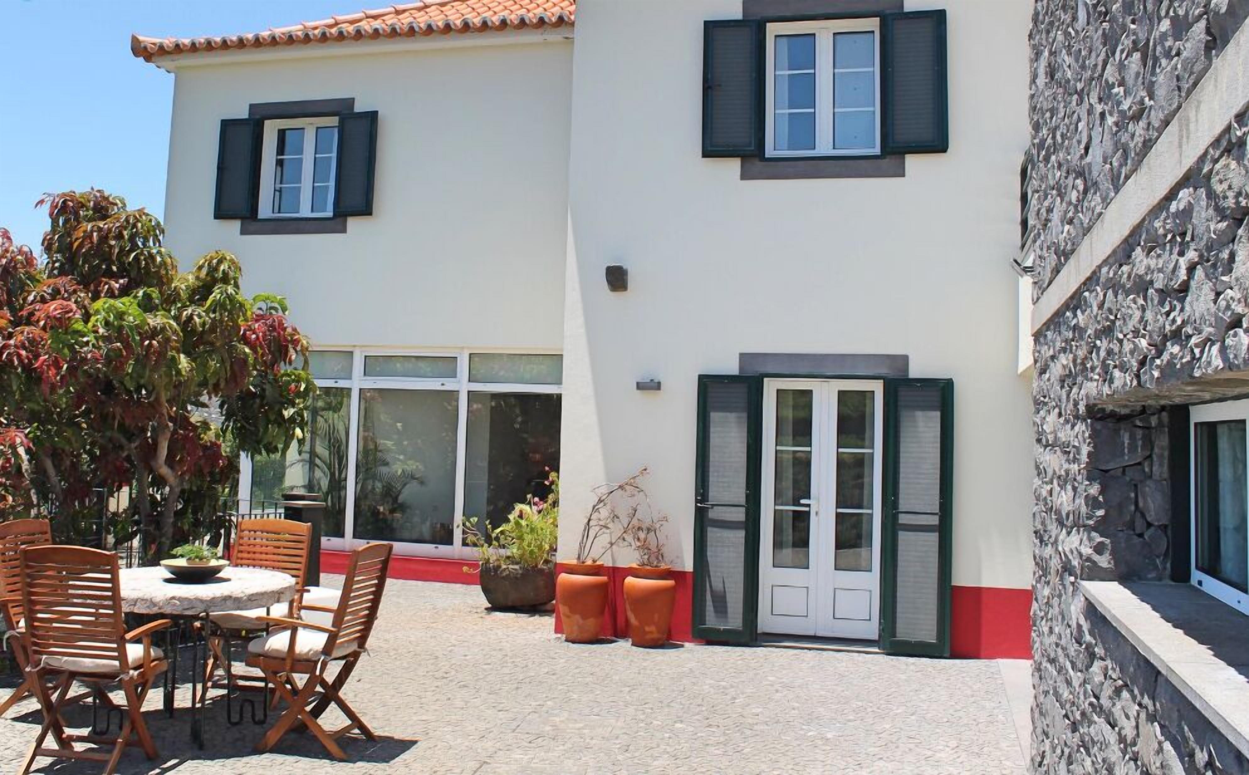 Rakennuksessa on tiilikatto, terassi, jossa on pyöreä pöytä ja tuoleja, sekä ruukkukasveja.