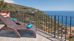 Ein Balkon mit zwei Liegestühlen und einem blauen Handtuch, mit Blick auf eine Küstenlandschaft mit Häusern und freiem Blick auf das Meer.