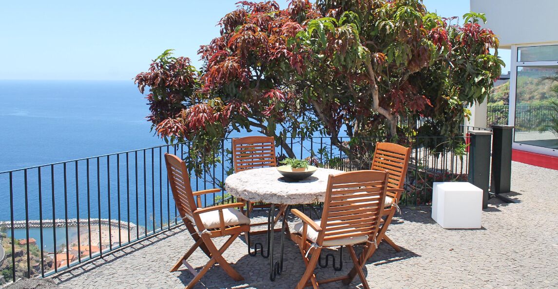 Una mesa redonda con cuatro sillas de madera, una planta en maceta y un cubo blanco en un balcón con vista al mar.