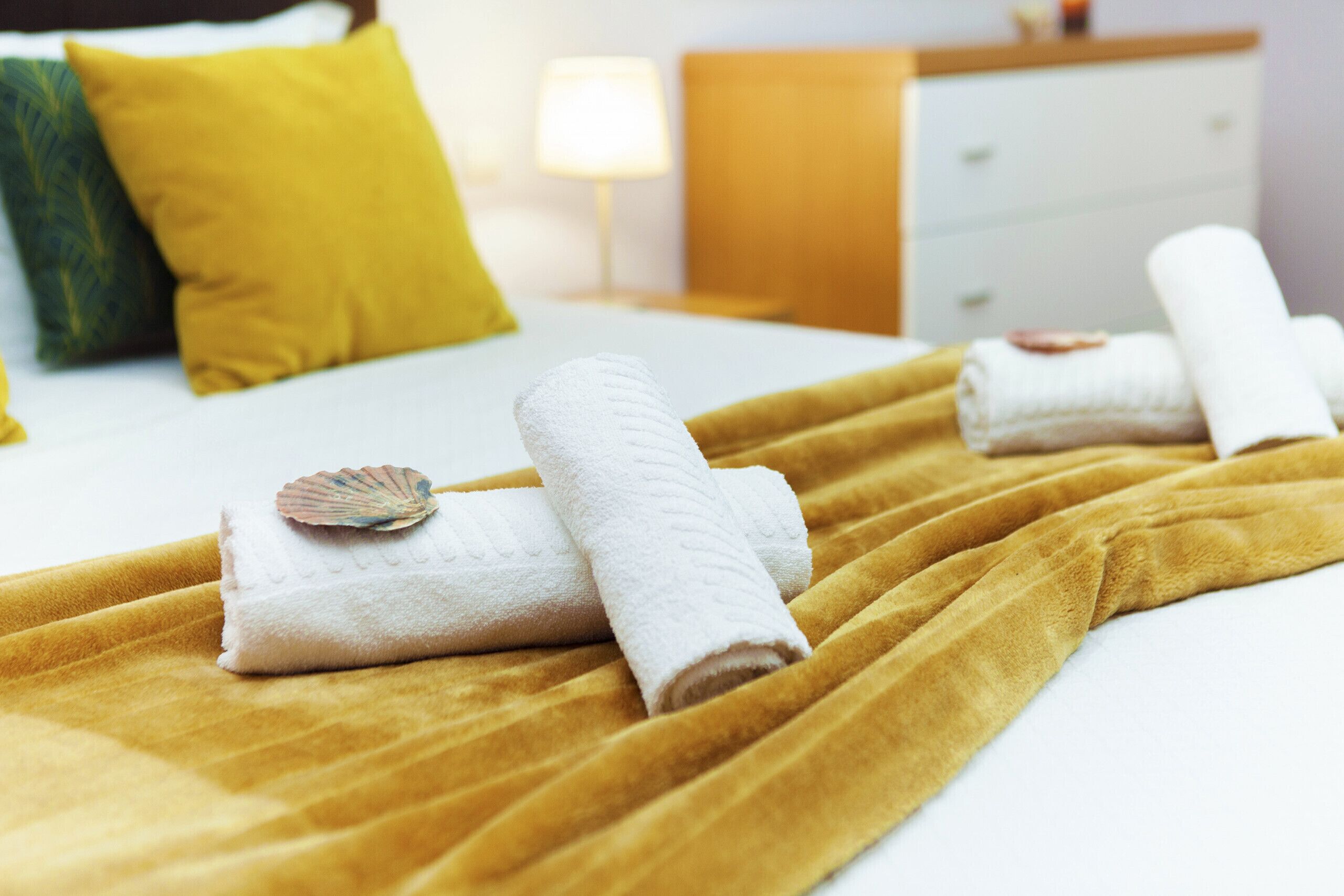 A neatly made bed with a yellow blanket, white towels, and a decorative shell.