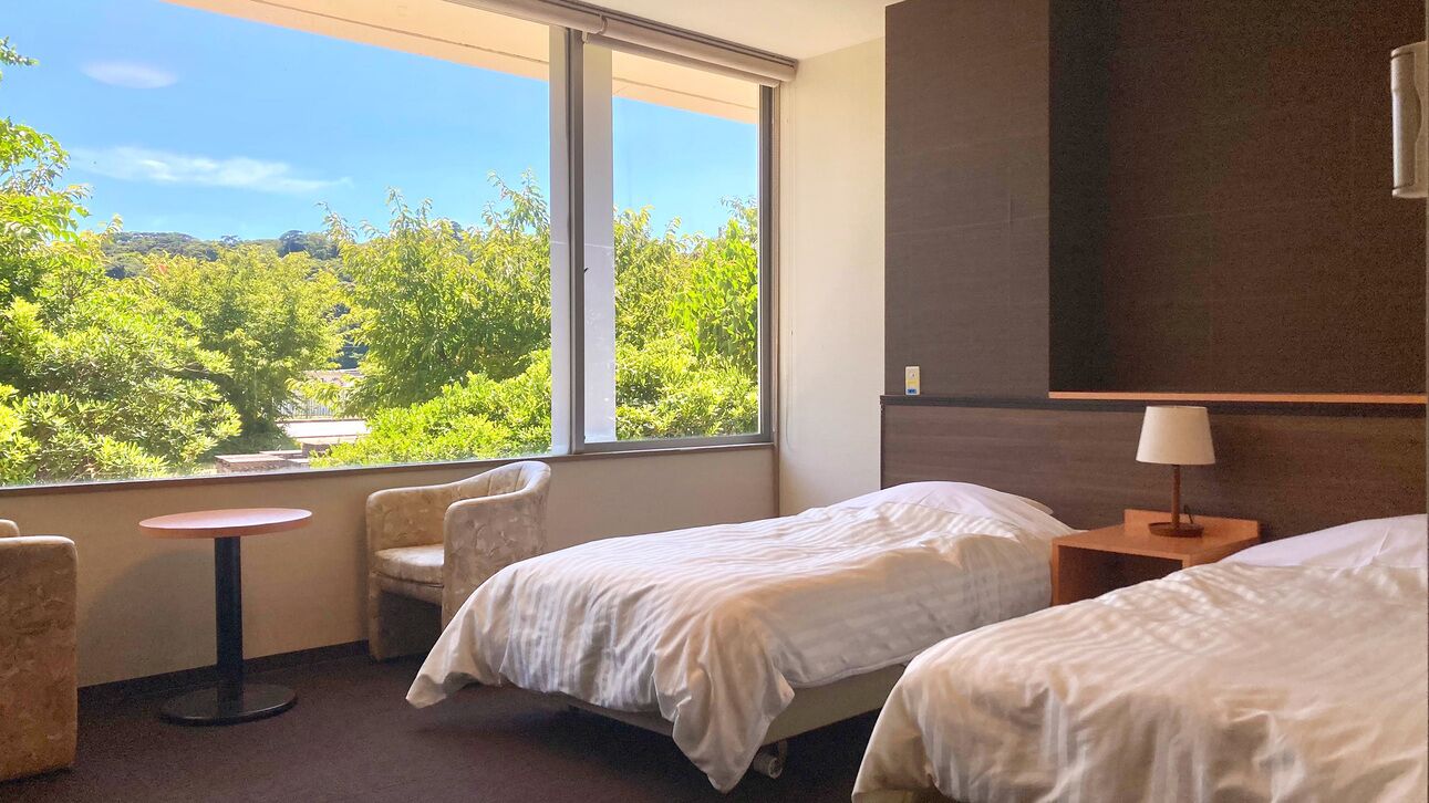 Hotelkamer met twee bedden, een klein rond tafeltje, een stoel, een lamp en een groot raam met uitzicht op groen.