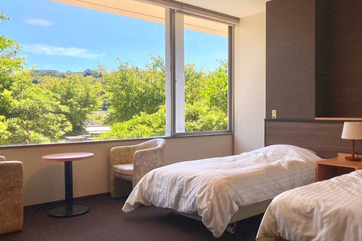 Habitación de hotel con dos camas, una mesa redonda pequeña, una silla, una lámpara y un ventanal con vistas a zonas verdes.