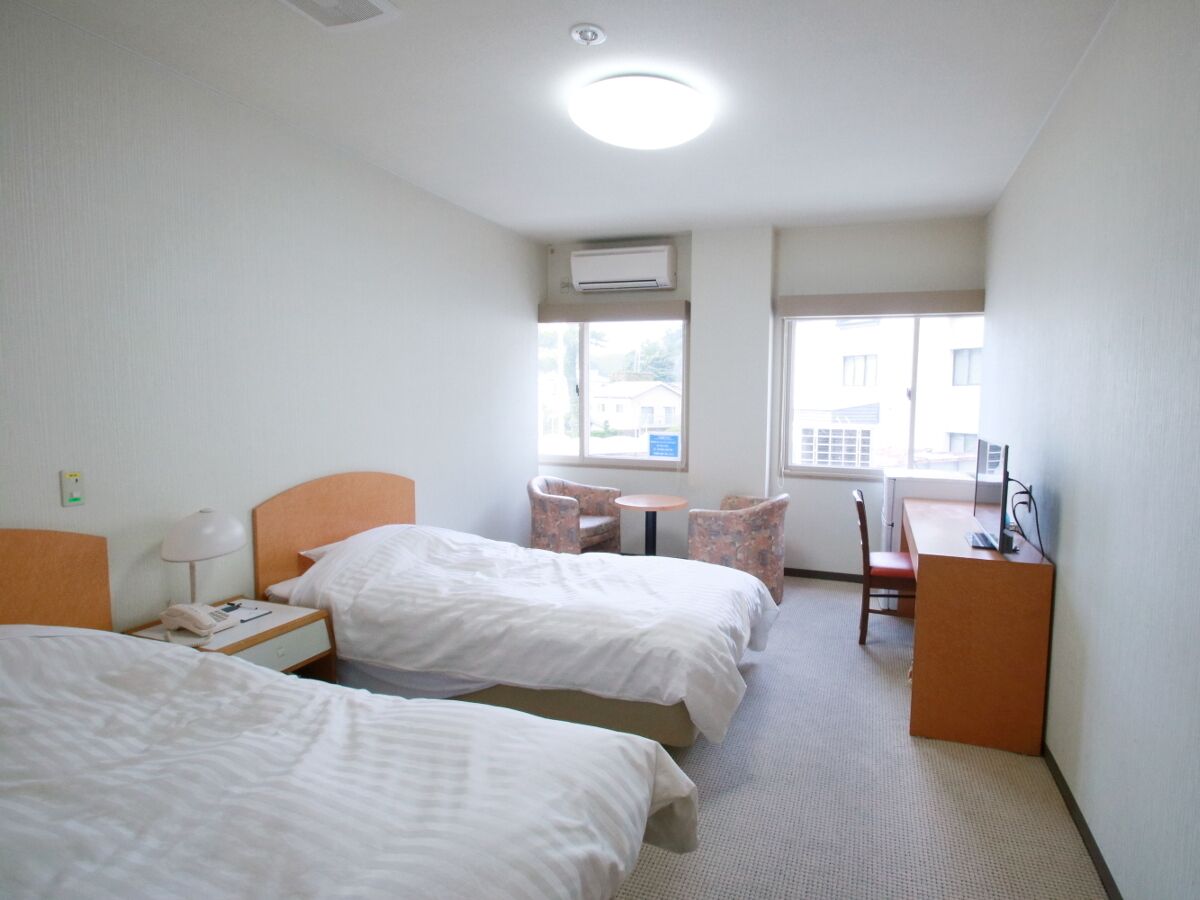 Ein Hotelzimmer mit zwei Betten, einem Schreibtisch, einem Stuhl, einem kleinen Tisch und einer Klimaanlage.