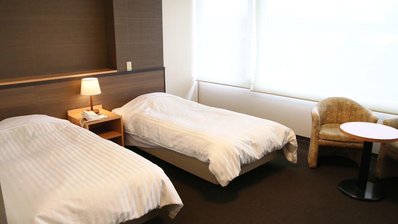 Een hotelkamer met twee eenpersoonsbedden, een nachtkastje met een lamp, een stoel en een klein rond tafeltje.