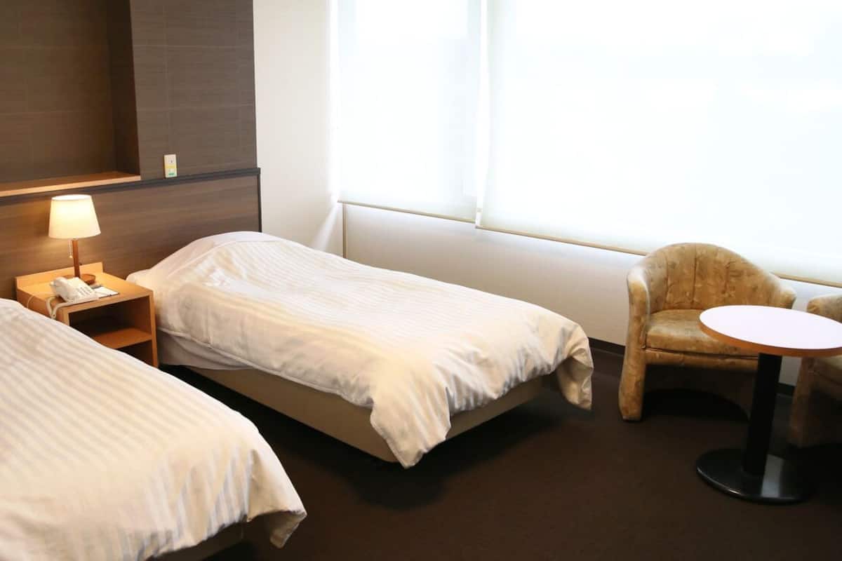 Habitación de hotel con dos camas individuales, una mesita de noche con lámpara, una silla y una pequeña mesa redonda.