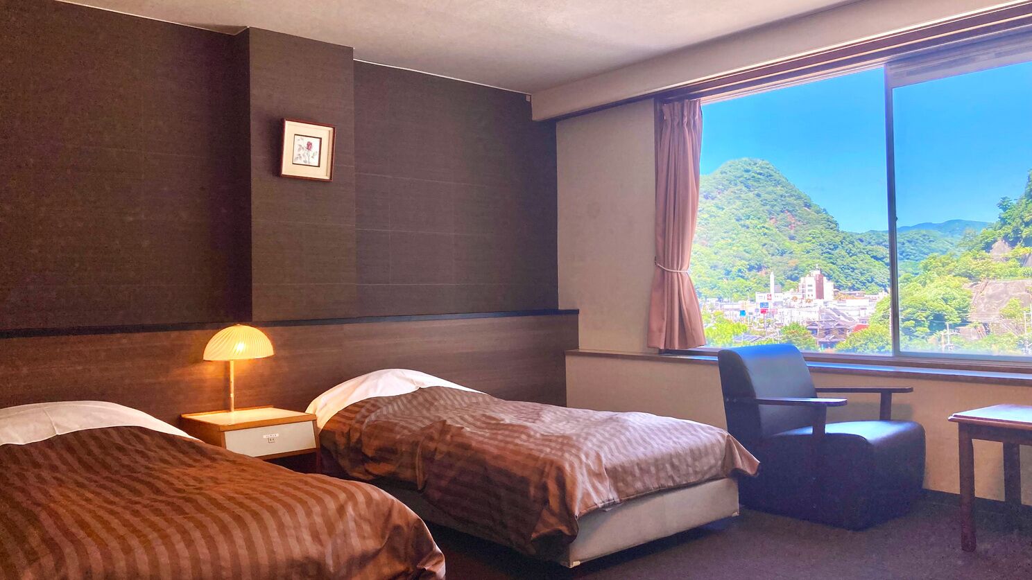 Hotelkamer met twee bedden, een bureau, een stoel en een groot raam met uitzicht op een bergachtig landschap.